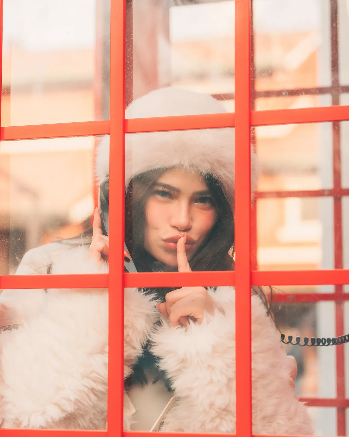 Denise Laurel behind the glass of a red telephone booth, finger to her lips