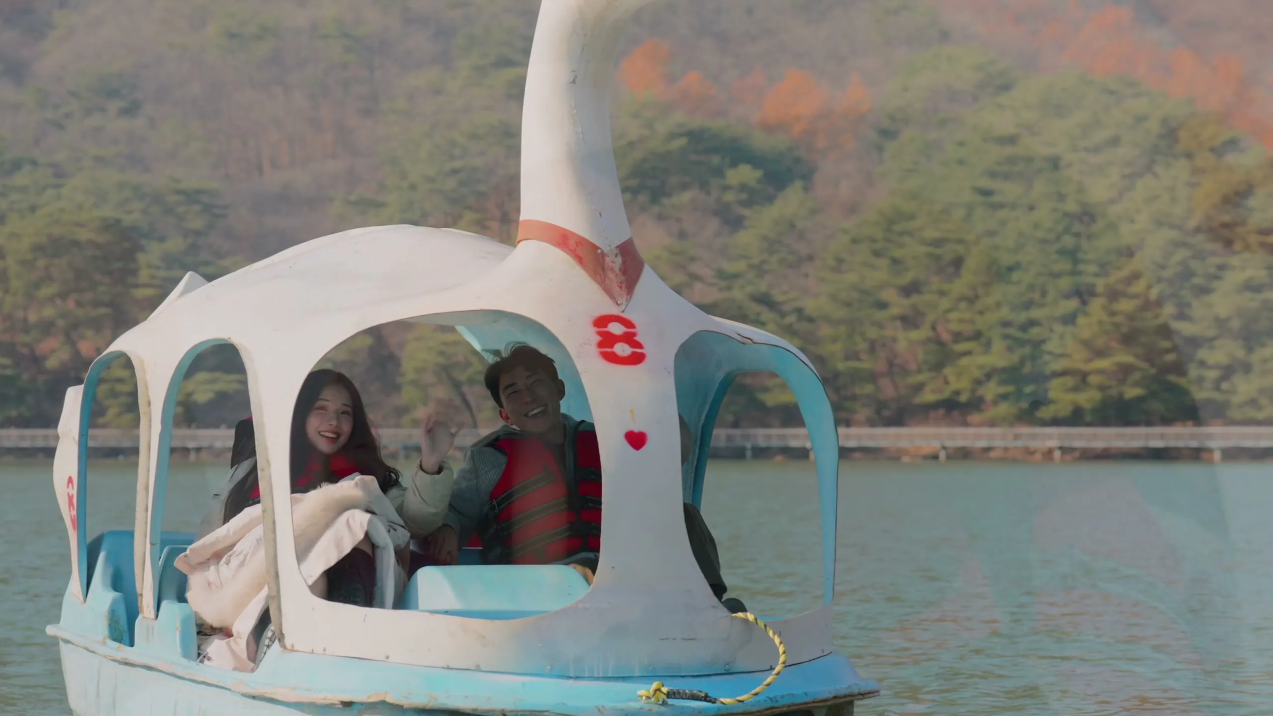 Out on the water in a swan pedal boat, the two of them waving from inside.