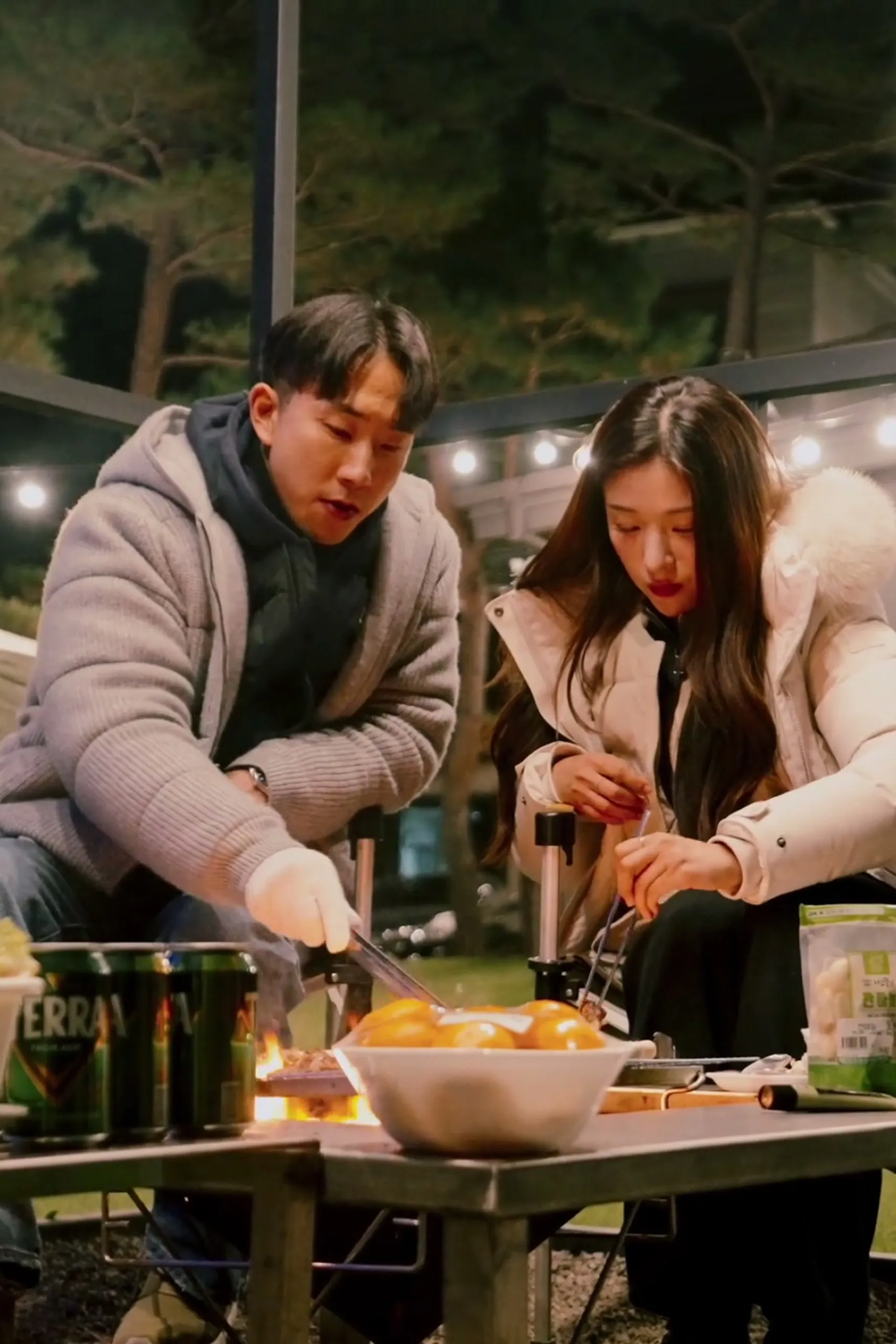 At the fire-lit table, the two of them grilling dinner together under the string lights.
