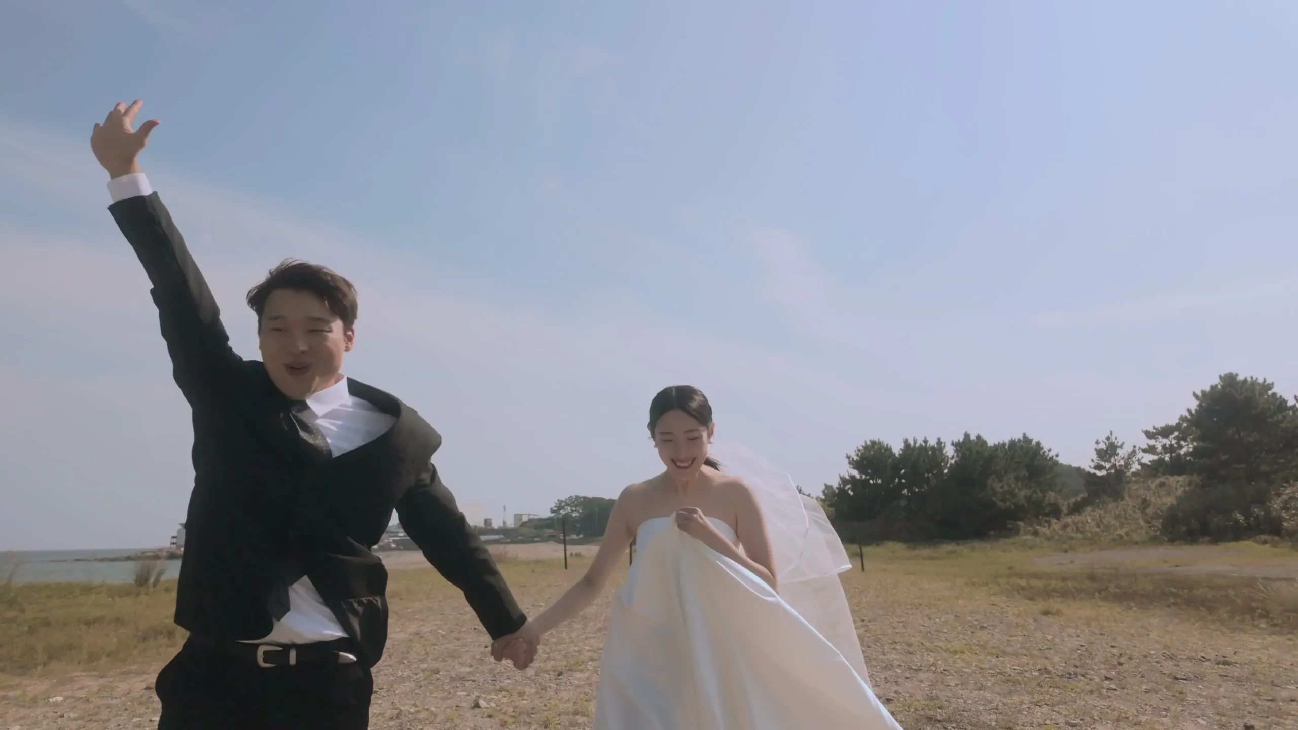In a field of autumn reeds, the groom throwing an arm up mid-step while the bride follows, both laughing.