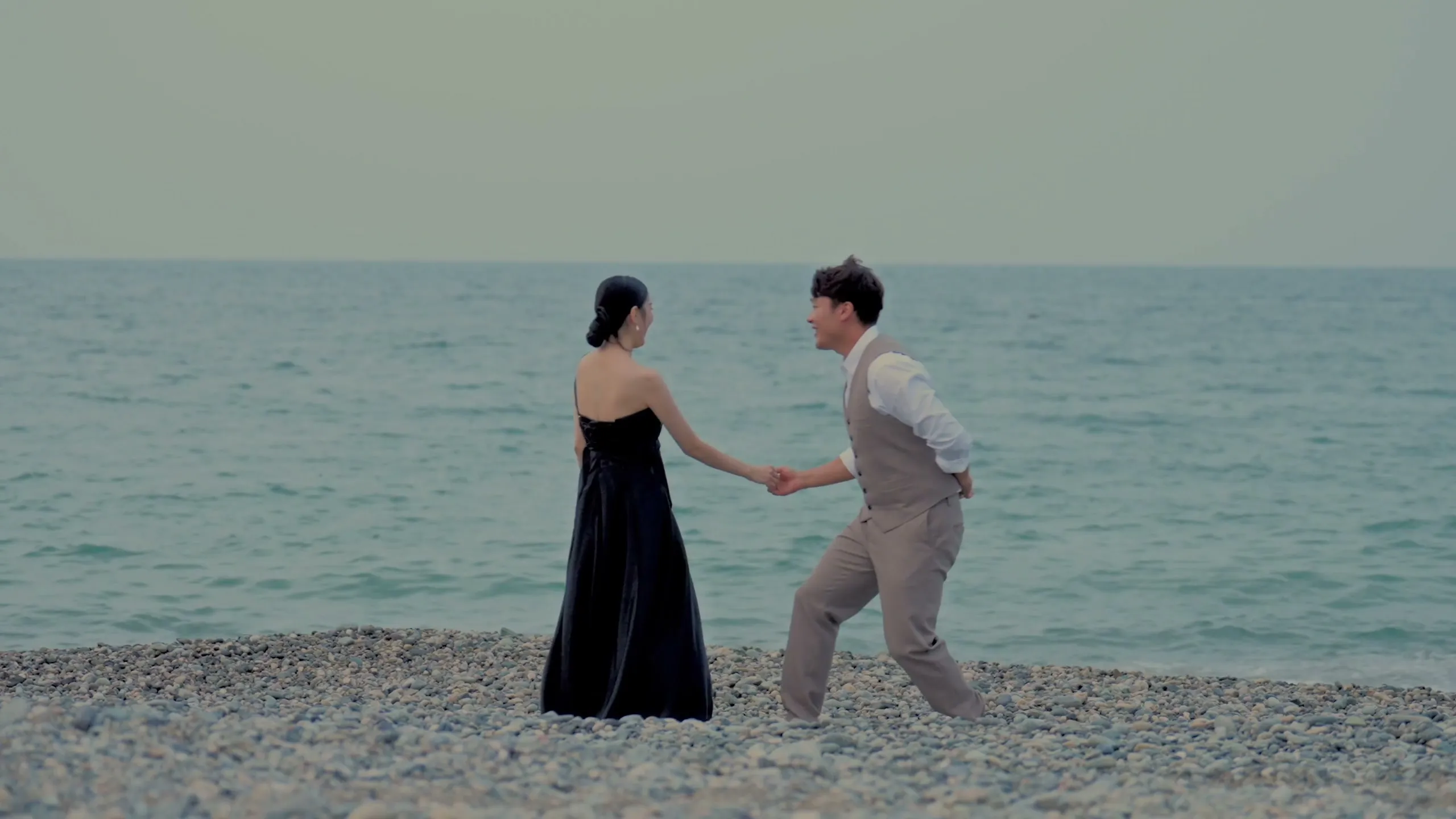 On the open pebble shore, the bride in a navy gown and the groom hand in hand, mid-stride by the sea.