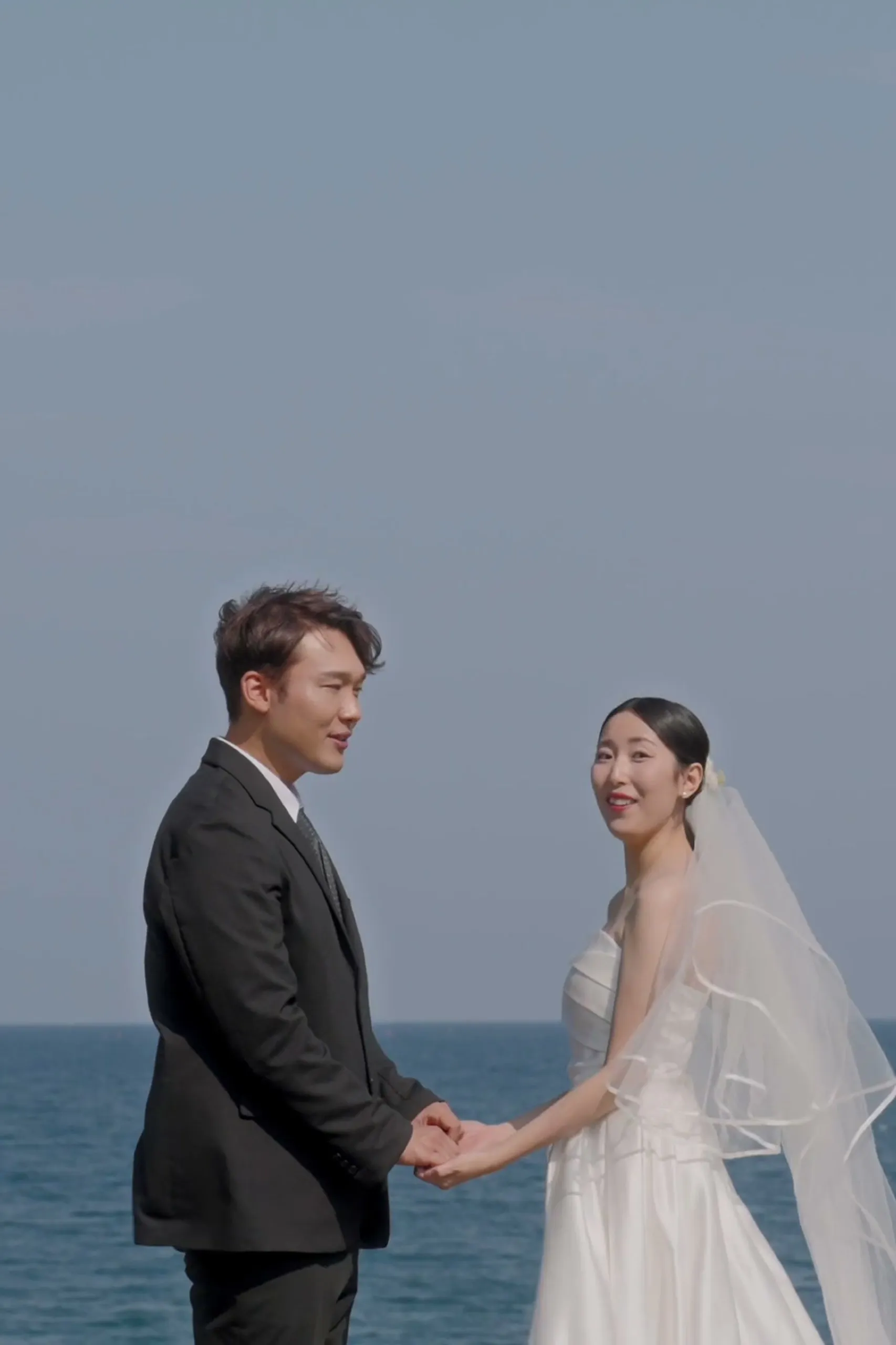 The two of them facing each other on the shore, the groom in a suit and the bride in her veil, the sea behind them.
