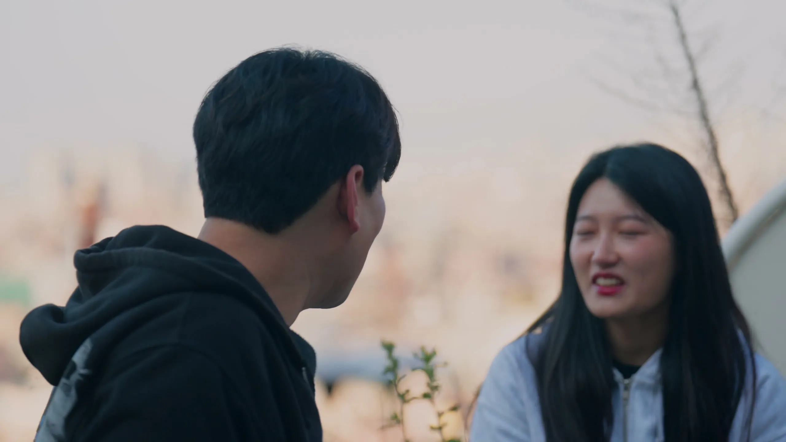 Haeseong and Chaeyeon walking down a sunlit Haebangchon street with the Seoul skyline behind them.