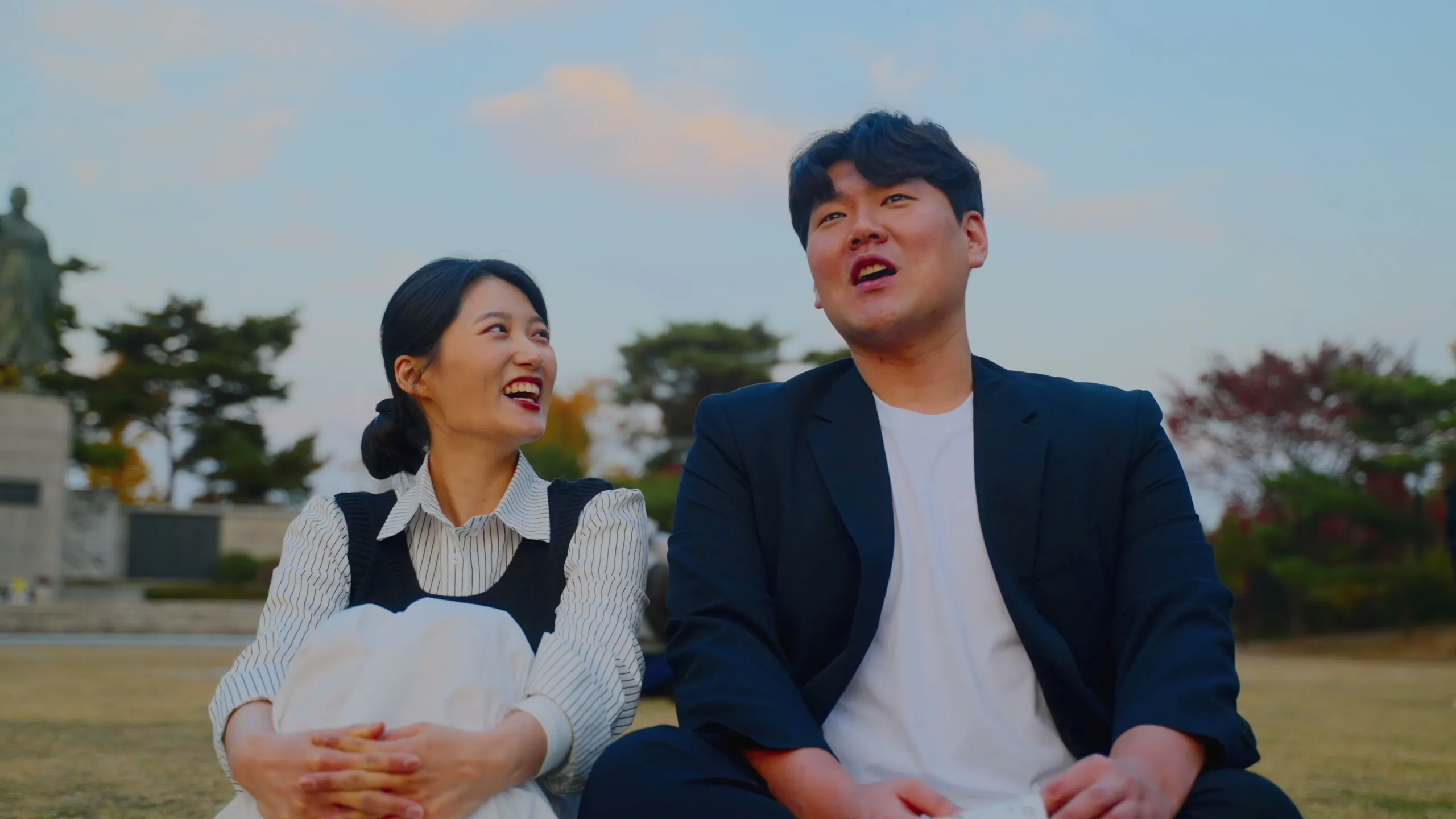 Haeseong and Chaeyeon walking down a sunlit Haebangchon street with the Seoul skyline behind them.