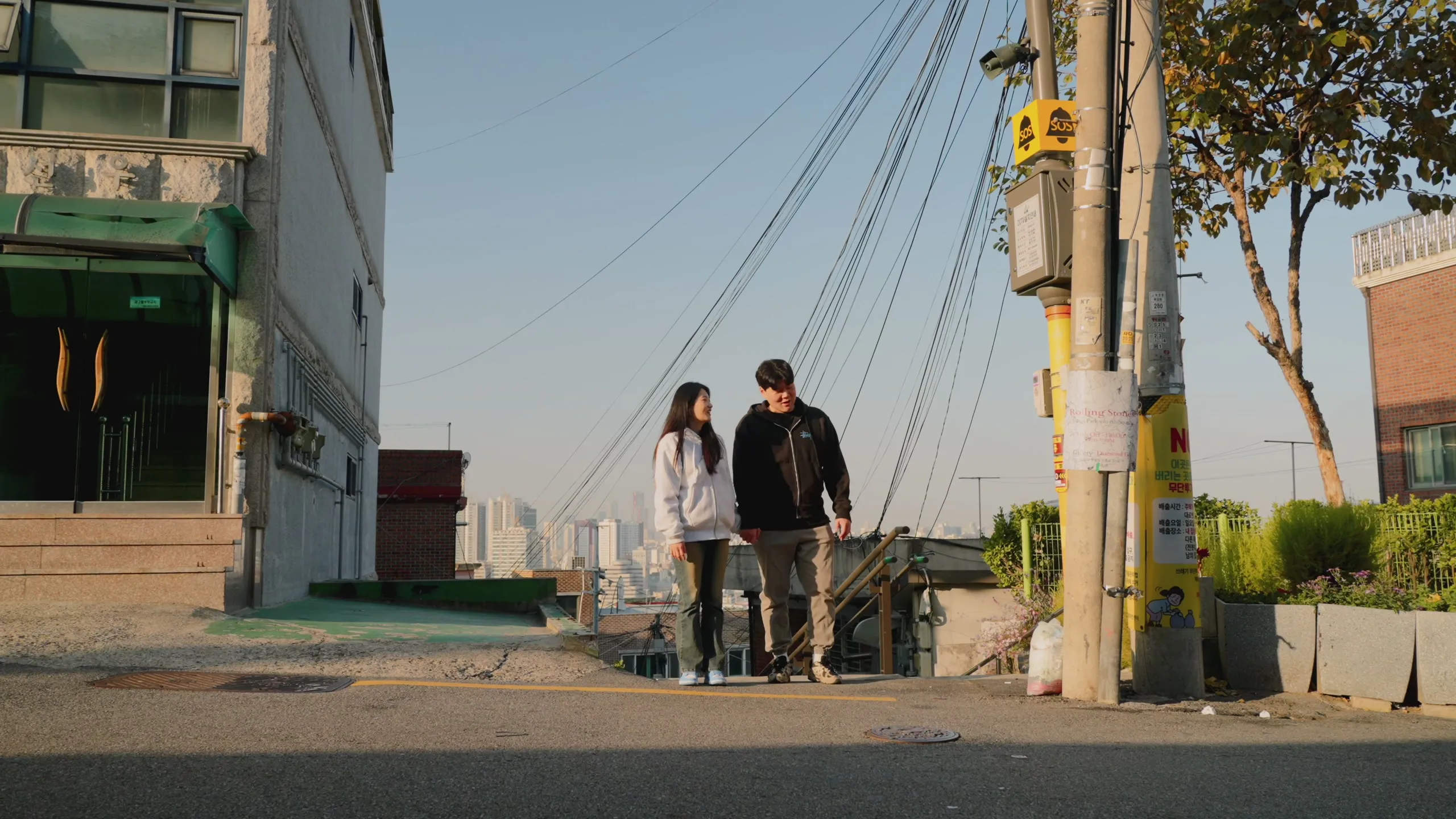 Haeseong and Chaeyeon walking a sunlit Haebangchon street with the Seoul skyline behind them