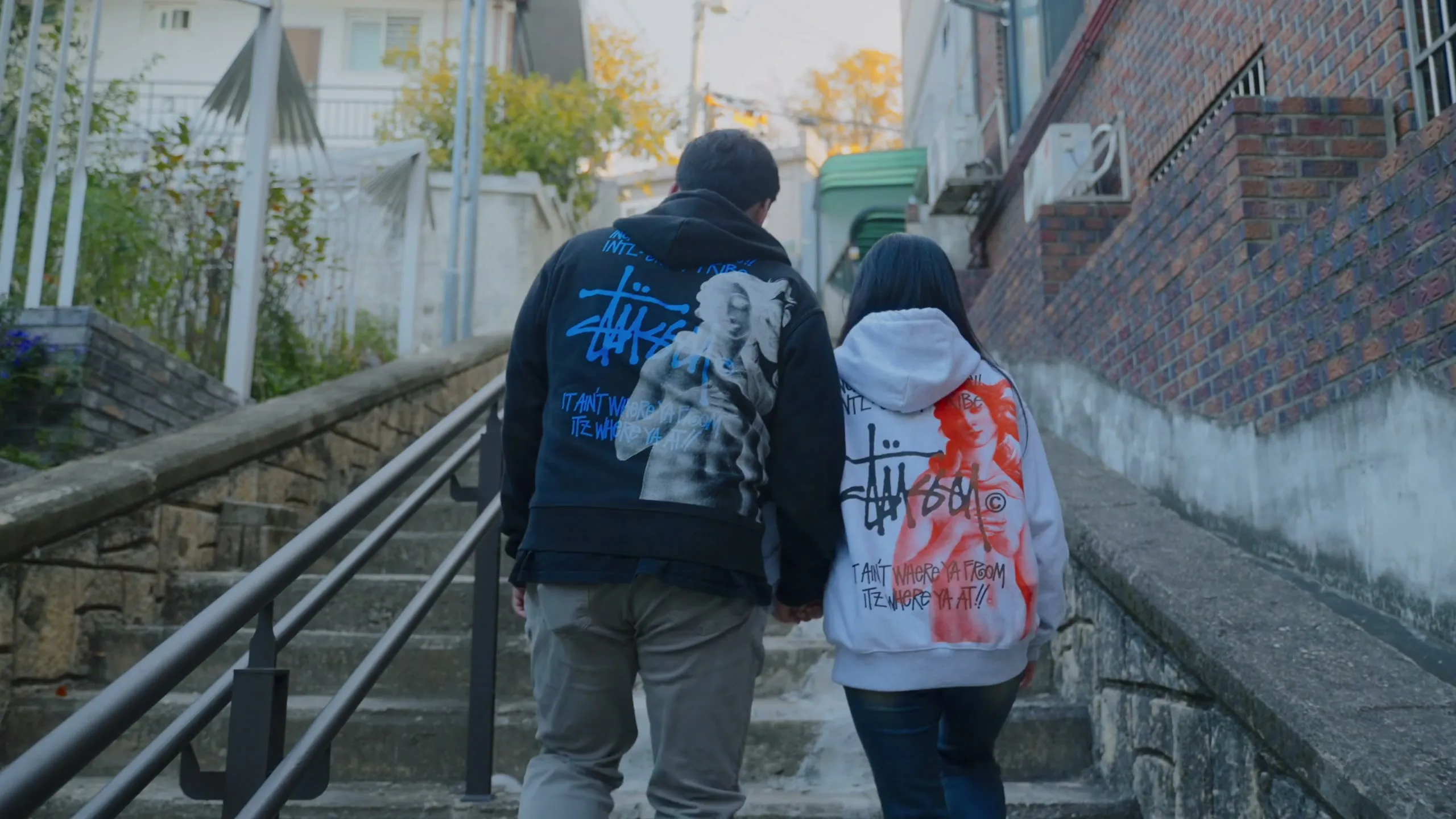Haeseong and Chaeyeon climbing a long flight of Haebangchon stairs, autumn leaves along the wall.
