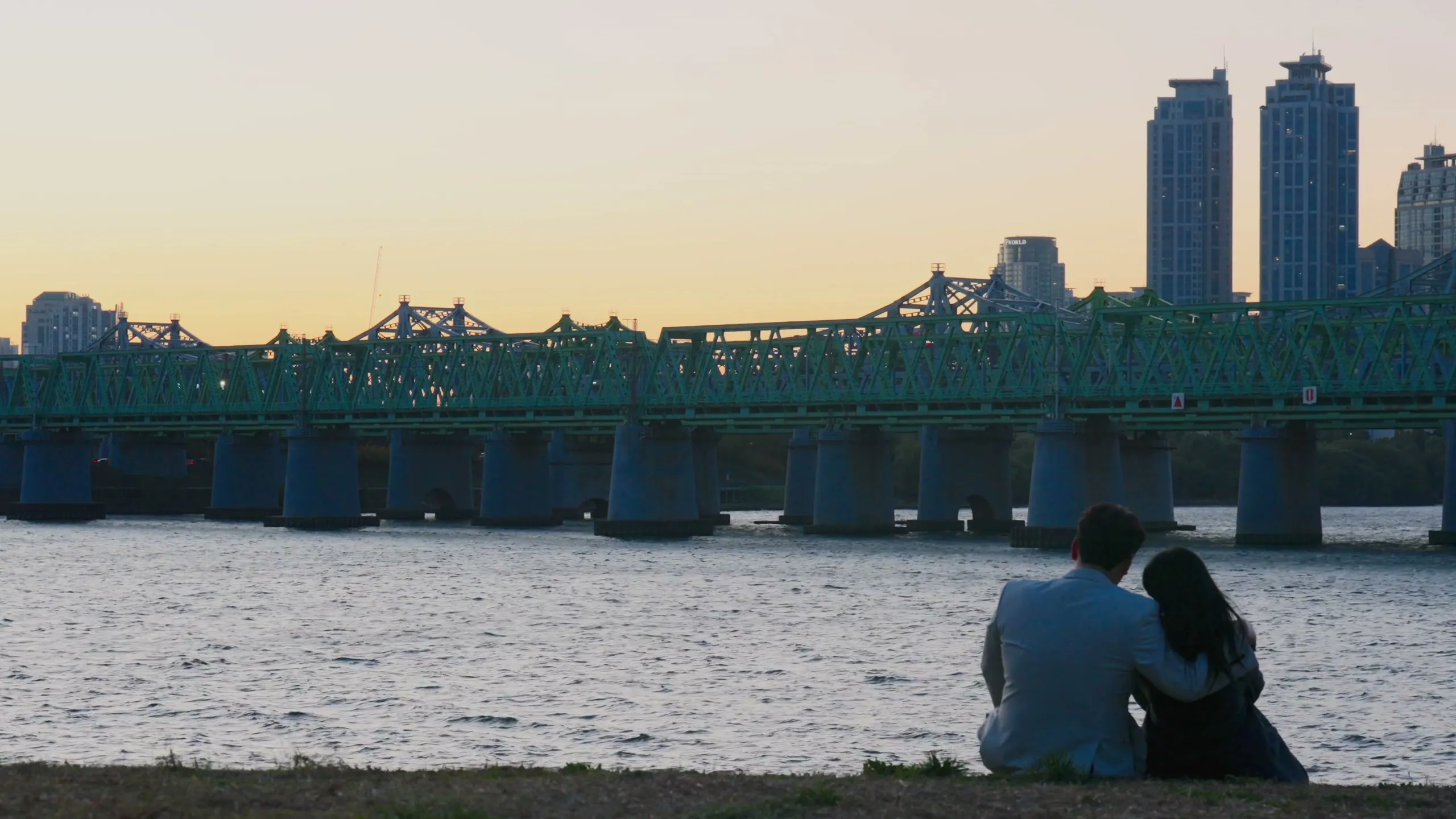 The couple small on the Han river bank at sunset, a bridge and the skyline stretched wide behind them.