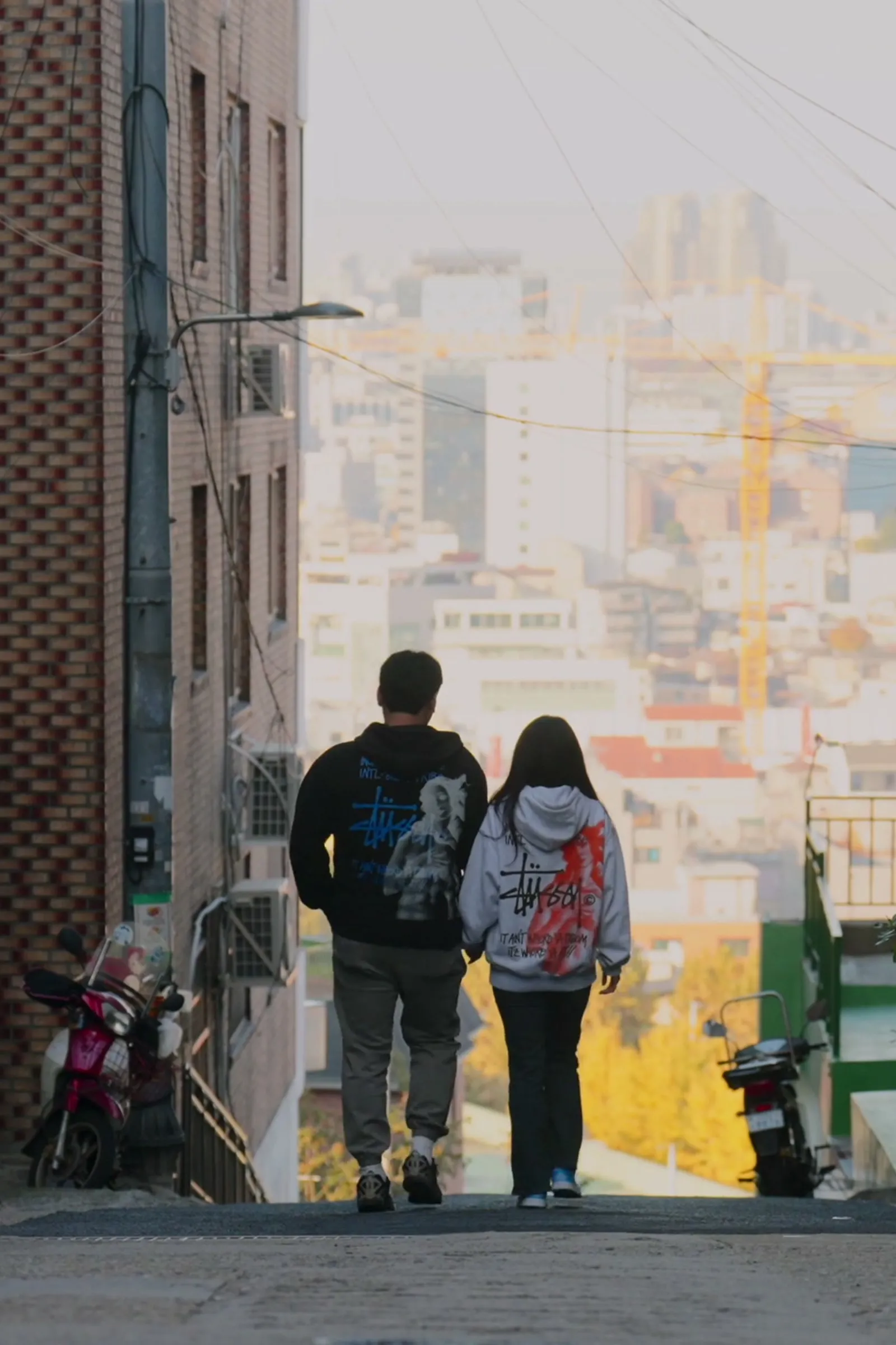 Haeseong and Chaeyeon walking away down a hillside alley toward the city view.