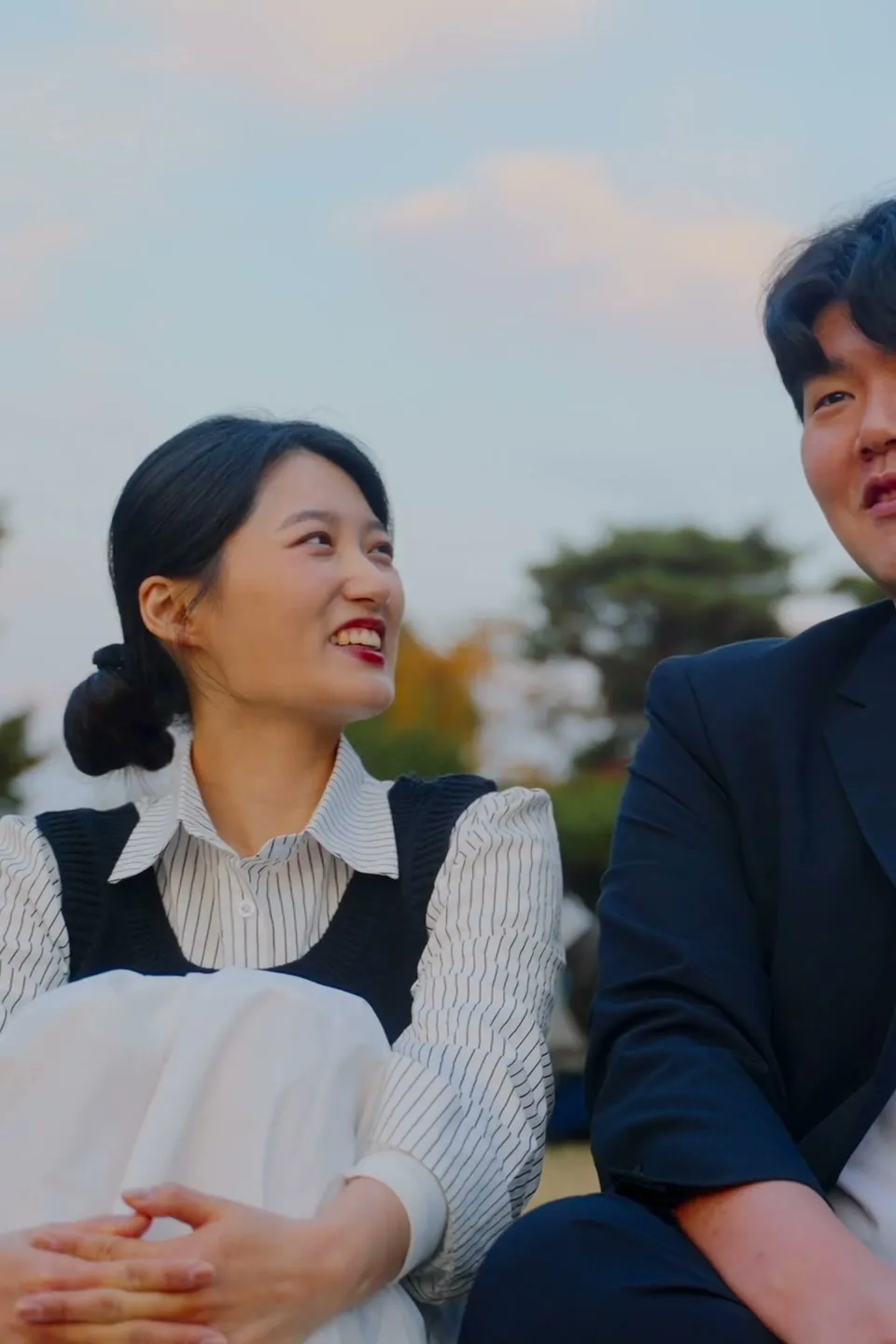Haeseong and Chaeyeon sitting close together on an autumn park lawn, dressed up for the day.